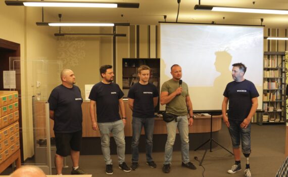 grupa prelegentów stoi przed ekranem projektora, jeden mówi do mikrofonu