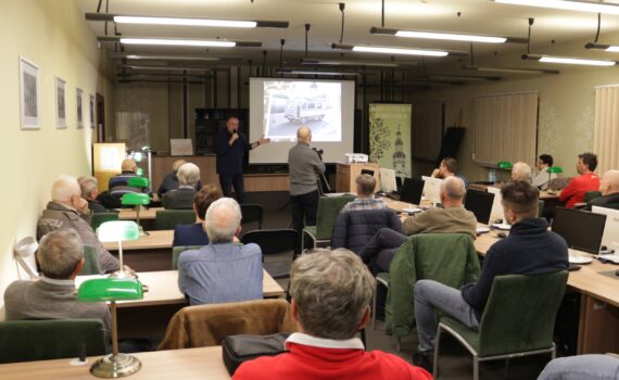 W jasnym pomieszczeniu z rzędami biurek i zielonymi lampkami odbywa się prezentacja. Na ekranie widoczne jest czarno-białe zdjęcie. Mężczyzna w czarnym golfie stoi przed ekranem i przemawia do grupy około 20 osób siedzących przy biurkach. Drugi mężczyzna stoi z boku, filmując prezentację.