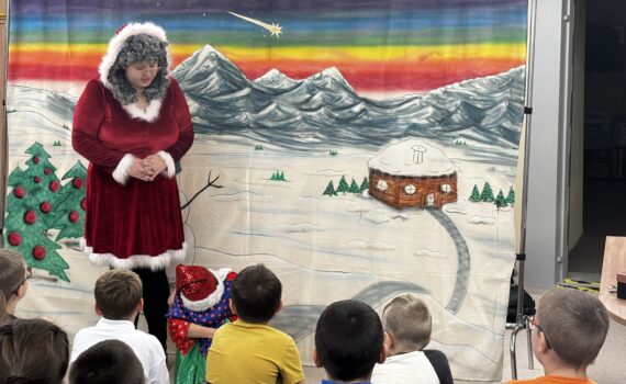 Mikołajka stoi na scenie. Dzieci siedzą na podłodze i patrzą na nich z zaciekawieniem.
