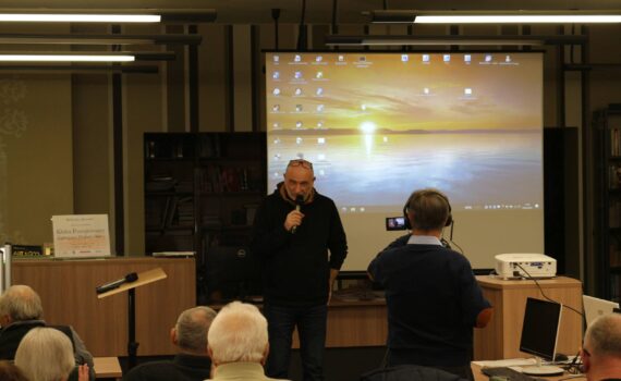 Na zdjęciu widzimy mężczyznę, który stoi przed grupą ludzi i trzyma mikrofon, przemawiając. Druga osoba, stojąca naprzeciwko mężczyzny nagrywa wydarzenie kamerą. W tle znajduje się duży ekran projekcyjny na którym wyświetlany jest pulpit z różnymi ikonami oraz tapetą przedstawiającą zachód słońca nad wodą. Po lewej stronie widoczny jest znak z napisem „Biblioteka 'Koronka'. Wygląda na to, że jest to biblioteka lub podobne miejsce, a wydarzenie to prezentacja lub wykład.