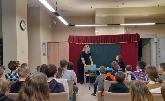 sala, prawdopodobnie w bibliotece lub szkole, w której odbywa się przedstawienie lub jakaś forma pokazu dla grupy dzieci. Na scenie, zaimprowizowanej za pomocą kurtyny, występują dwie lub trzy osoby. Widzowie siedzą na krzesłach zwróceni w stronę sceny. Jedna z aktorek jest ubrana w biały strój, prawdopodobnie kostium, i wykonuje jakąś czynność przy stole. Druga aktorka siedzi przy stole. Wygląda to na konkretną scenę z przedstawienia.