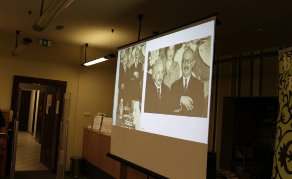 Zdjęcie przedstawia projekcję na ekranie. Widoczne jest stare, czarno-białe zdjęcie grupowe. Na pierwszym planie znajdują się dwaj mężczyźni w garniturach, jeden z nich przypomina Alberta Einsteina. W tle widoczne są inne osoby, prawdopodobnie kobiety w strojach z epoki. Na dole zdjęcia widnieje rok "1930". Całość sugeruje prezentację historycznych fotografii.