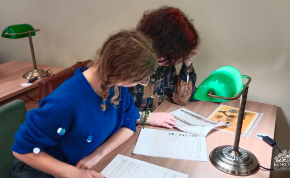 Opis wygenerowany automatycznie: Na tym zdjęciu widzimy dwie młode kobiety, pochłonięte lekturą. Siedzą przy biurku, na którym stoi zielona lampa, podobnie jak na pierwszym zdjęciu. Scena jest kameralna i skupiona na akcie czytania. Delikatne światło lampy tworzy intymną atmosferę, podkreślając spokój i skupienie kobiet. Uwagę przyciąga spokój i skupienie obu kobiet.