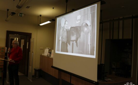 Opis wygenerowany automatycznie: zdjęcie przedstawia projekcję czarno-białego filmu na ekranie. Na filmie widać mężczyznę, który trzyma w ręku duży aparat fotograficzny. W tle widać pomieszczenie biblioteki.