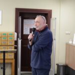 Opis wygenerowany automatycznie: Zdjęcie przedstawia mężczyznę w okularach przemawiającego do mikrofonu. Ubrany jest w ciemną, jednokolorową bluzę z długim rękawem. Tło stanowi wnętrze biblioteki, widoczne są drewniane regały z katalogiem kartotekowym. Mężczyzna stoi za niewielkim, drewnianym pulpitem. Całość kompozycji sugeruje formalne wystąpienie publiczne w akademickim lub bibliotecznym otoczeniu.