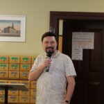 Opis wygenerowany automatycznie: Zdjęcie przedstawia mężczyznę przemawiającego do mikrofonu. Ubrany jest we wzorzystą koszulę. Tło stanowi wnętrze biblioteki, widoczne są drewniane regały z katalogiem kartotekowym. Mężczyzna stoi za niewielkim, drewnianym pulpitem. Całość kompozycji sugeruje formalne wystąpienie publiczne w akademickim lub bibliotecznym otoczeniu.