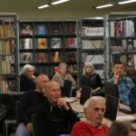 Opis wygenerowany automatycznie: Zdjęcie przedstawia grupę osób siedzących w bibliotece. Osoby te skupiają się na czymś, co dzieje się przed nimi, prawdopodobnie na prezentacji lub spotkaniu. W tle widoczne są regały z książkami i czasopismami.