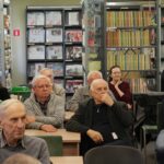 Opis wygenerowany automatycznie: Zdjęcie przedstawia kameralną grupę osób siedzących w bibliotece. Oświetlenie jest ciepłe i miękkie, tworząc spokojną atmosferę. Osoby na zdjęciu są skupione, pochłonięte słuchaniem czegoś – być może wykładu, opowieści, lub dyskusji. Ich mimika sugeruje zainteresowanie i zaangażowanie. Widoczne są książki na półkach w tle, podkreślające kontekst biblioteczny.