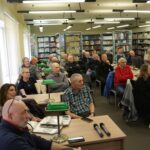 Opis wygenerowany automatycznie: To zdjęcie przedstawia grupę osób w większym i bardziej nowoczesnym pomieszczeniu. W tle widoczne są komputery, sugerujące, że grupa może brać udział w warsztatach komputerowych lub korzystając z technologii cyfrowej w celach edukacyjnych lub społecznościowych. Atmosfera wydaje się być skupiona ale bardziej dynamiczna.