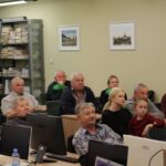 Opis wygenerowany automatycznie: Zdjęcie przedstawia grupę osób siedzących w sali wykładowej. Widoczne są komputery, a w tle regały z książkami. Osoby na zdjęciu wyglądają na skupione i uważnie słuchające.