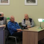 Opis wygenerowany automatycznie: Zdjęcie pokazuje mniejszą grupę osób siedzących przy stole. Na stole stoi lampa i znajduje się jakiś przedmiot, prawdopodobnie książka. Tło jest stonowane, w jasnych kolorach. Atmosfera wydaje się być bardziej kameralna.