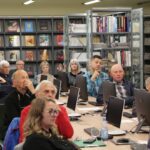 Opis wygenerowany automatycznie: Zdjęcie przedstawia grupę osób siedzących przy komputerach w sali wykładowej. Tło to regały wypełnione książkami. Osoby na zdjęciu wydają się być zaangażowane w to, co się dzieje.