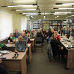 Opis wygenerowany automatycznie: Sala wykładowa wypełniona słuchaczami. Widok sugeruje akademickie środowisko, być może uniwersytet lub szkołę wyższą. Słuchacze siedzą skupieni, sugerując zaangażowanie w trwającą prezentację lub wykład. Atmosfera wydaje się być skupiona i poważna. Można zauważyć różnorodność słuchaczy pod względem wieku i ubioru.