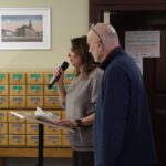 Opis wygenerowany automatycznie: Kobieta z mikrofonem przemawiająca do publiczności. Jej postawa jest pewna i profesjonalna, sugerując doświadczenie w wystąpieniach publicznych. Wyraźnie widać, że skupia uwagę zebranych. Mikrofon wskazuje na formalny charakter wydarzenia, być może konferencję, seminarium lub spotkanie.
