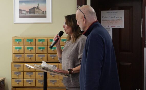 Opis wygenerowany automatycznie: Kobieta z mikrofonem przemawiająca do publiczności. Jej postawa jest pewna i profesjonalna, sugerując doświadczenie w wystąpieniach publicznych. Wyraźnie widać, że skupia uwagę zebranych. Mikrofon wskazuje na formalny charakter wydarzenia, być może konferencję, seminarium lub spotkanie.