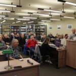 Opis wygenerowany automatycznie: Zdjęcie przedstawia salę biblioteczną wypełnioną słuchaczami skupionymi na prelegencie. Widzimy osoby w różnym wieku, siedzące przy stołach z komputerami. W tle znajdują się regały wypełnione książkami. Na pierwszym planie stoi stół z mikrofonem, a na końcu sali stoi prelegent przy projektorze. Atmosfera jest skupiona i formalna, sugerująca wykład, prezentację lub warsztat. Oświetlenie jest jasne, a kolorystyka stonowana.