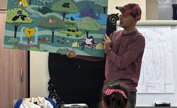 Opis wygenerowany automatycznie: Zdjęcie przedstawia mężczyznę pokazującego dzieciom kolorową ilustrację. Rysunek jest bogaty w detale: widzimy góry, drzewa, różne zwierzęta i samochód. Mężczyzna z uśmiechem wyjaśnia dzieciom szczegóły ilustracji, angażując je w proces poznawania.