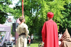 Opis wygenerowany automatycznie: Ci sami uczestnicy inscenizacji, tym razem stojący. W tle widoczny miecz, sugerujący kontekst historyczny lub legendę. Kompozycja zdjęcia podkreśla napięcie i oczekiwanie.