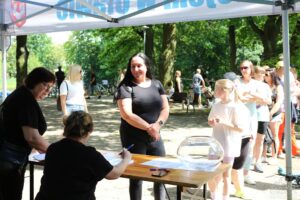 Opis wygenerowany automatycznie: Stoisko informacyjne lub rejestracyjne na imprezie plenerowej. Kilka osób stoi w kolejce, a wolontariuszki obsługują uczestników. W tle widać park i innych ludzi.