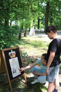 Opis wygenerowany automatycznie: Kobieta pokazuje chłopcu tablicę z informacjami. Tablica ma napis "Pierścionek i korona". W tle widać drzewa i ścieżkę.