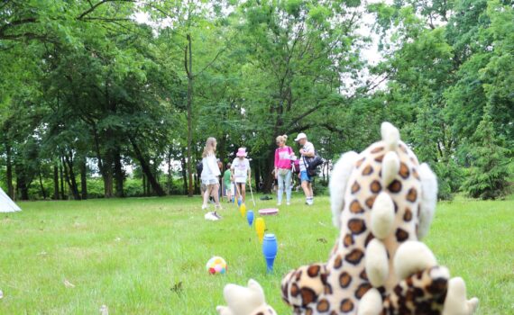 Opis wygenerowany automatycznie: Wesoła grupa dzieci i dorosłych bawi się na trawiastej polanie. Na pierwszym planie znajduje się pluszowa żyrafa. Scena emanuje radością i beztroską, sugerując wspólne spędzanie czasu na świeżym powietrzu.