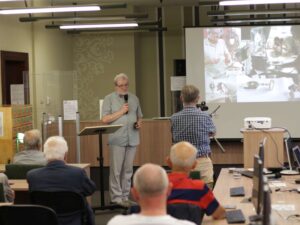 Opis wygenerowany automatycznie: Mężczyzna wygłasza prezentację na konferencji. Na ekranie za nim widoczne są slajdy, a publiczność siedzi przy stołach z laptopami.