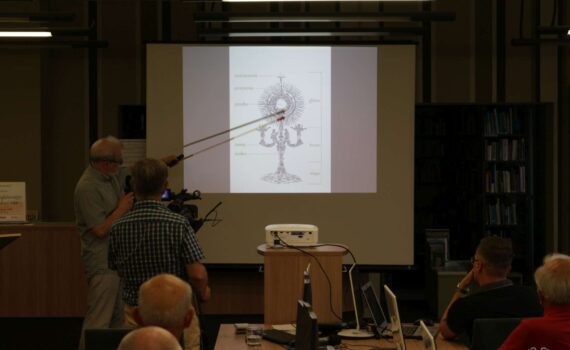 Opis wygenerowany automatycznie: Prelegent, wskazując laską na ekran, kontynuuje prezentację. Sala konferencyjna wypełniona jest słuchaczami skupionymi na wystąpieniu.