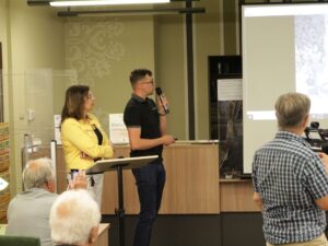 Opis wygenerowany automatycznie: Mężczyzna i kobieta stoją przy podium i przemawiają do publiczności. Na ekranie za nimi wyświetlane jest zdjęcie. W tle widoczna jest część biblioteki.
