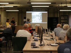 Opis wygenerowany automatycznie: Mężczyzna i kobieta stoją przy podium i przemawiają do publiczności. Na ekranie za nimi wyświetlane jest zdjęcie. W tle widoczna jest część biblioteki.
