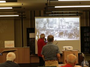 Opis wygenerowany automatycznie: Prezentacja historyczna. Mężczyzna wskazuje na czarno-białe zdjęcie ulicy z grupą ludzi z epoki. Publiczność uważnie słucha.