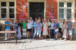 Grupa ludzi pozuje do zdjęcia przed budynkiem o pomarańczowych ścianach i dużych, ciemnych drzwiach. Na ścianie widnieją napisy: "Rada Miejska w Oławie" oraz "Powiatowa i Miejska Biblioteka Publiczna w Oławie". Niektórzy z obecnych trzymają niebieski transparent z napisem "EDD". Teren przed budynkiem wyłożony jest brukiem. Cała scena sugeruje wydarzenie o charakterze publicznym związane z biblioteką, z wyraźnym akcentem na inicjatywę "EDD".