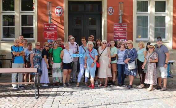 Grupa ludzi pozuje do zdjęcia przed budynkiem o pomarańczowych ścianach i dużych, ciemnych drzwiach. Na ścianie widnieją napisy: "Rada Miejska w Oławie" oraz "Powiatowa i Miejska Biblioteka Publiczna w Oławie". Niektórzy z obecnych trzymają niebieski transparent z napisem "EDD". Teren przed budynkiem wyłożony jest brukiem. Cała scena sugeruje wydarzenie o charakterze publicznym związane z biblioteką, z wyraźnym akcentem na inicjatywę "EDD".