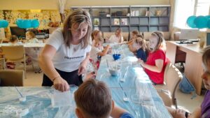Kobieta w białej koszulce z napisem "@Gipsoweckzi" przelewa płyn z pojemnika do kubka na stole przykrytym niebieską ceratą. Wokół stołu siedzi kilkoro dzieci, zaangażowanych w jakąś aktywność. W tle widać niebieskie balony.