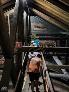 Ludzie spacerują po drewnianej kładce wewnątrz ciemnego, industrialnego pomieszczenia z widocznymi drewnianymi belkami i rurami. Na jednej z drewnianych belek widoczny jest czerwony znak gaśnicy.