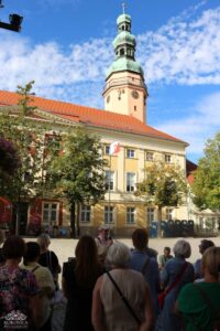 Grupa ludzi zebrała się na placu miejskim, słuchając przemawiającego mężczyzny. Za nimi znajduje się budynek z wieżą zegarową i dachem pokrytym czerwonymi dachówkami. Niebo jest błękitne z kilkoma chmurami.