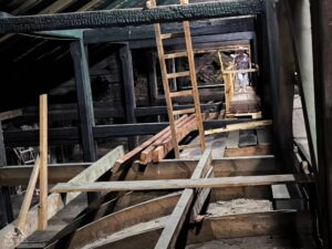 Grupa osób znajduje się w słabo oświetlonym pomieszczeniu na poddaszu, z widocznymi drewnianymi belkami stropowymi i drabiną.