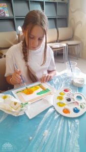 Dziewczynka z warkoczem maluje obrazek żółtymi i pomarańczowymi farbami przy stole z paletą i pędzlami.