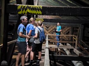Grupa ludzi słucha przewodnika na drewnianym strychu z widocznymi belkami i rurami.