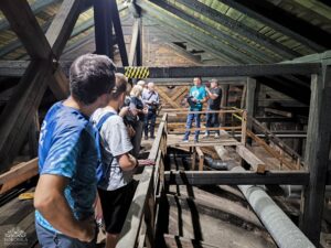 Grupa ludzi słucha przewodnika na drewnianym strychu z widocznymi belkami i rurami.