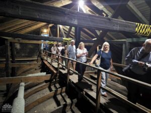 Grupa osób przemierza drewnianą kładkę, która prowadzi przez wnętrze strychu. Wokół nich widoczne są surowe, odsłonięte belki stropowe oraz elementy konstrukcyjne dachu, tworząc surowy i historyczny klimat przestrzeni.