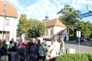 Grupa osób stoi na rogu ulicy, wpatrując się w niewielki budynek z wieżyczką. Na znaku ulicznym widnieje napis "ul. Św. Rocha". Pogoda jest słoneczna z kilkoma chmurami na niebie.