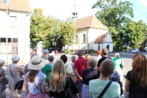 Grupa ludzi stoi tyłem do widza, obserwując mały, jasny budynek z czerwonym dachem i wieżyczką, otoczony drzewami.