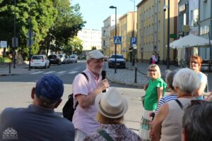 Mężczyzna w charakterystycznej różowej koszuli i czapce przemawia do zebranej na ulicy grupy osób.