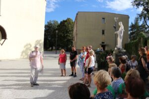 Na tle placu z okazałym posągiem, mężczyzna w czapce i różowej koszuli prowadzi rozmowę z grupą słuchaczy.