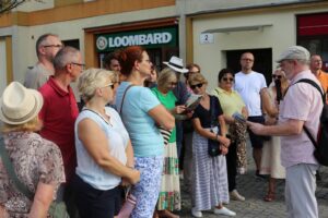 Przed witryną sklepu z wyraźnym napisem "LOOMBARD", mężczyzna ubrany w czapkę i różową koszulę zwraca uwagę grupy ludzi.