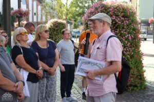 Mężczyzna w różowej koszuli i czapce, trzymający mikrofon i dokument, przemawia do grupy uważnie słuchających osób. W tle rozpościera się duży krzew obsypany różowymi kwiatami, tworząc malowniczą scenerię dla tego wydarzenia.