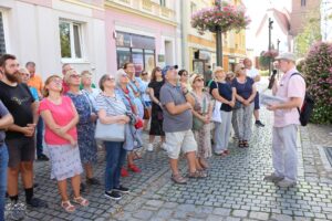 Grupa ludzi słucha mężczyzny z mikrofonem na zewnątrz. Mężczyzna trzyma papiery i wydaje się prowadzić wycieczkę lub prezentację. W grupie są mężczyźni i kobiety w różnym wieku, wszyscy patrzą w tym samym kierunku, prawdopodobnie na coś, na co wskazuje lub o czym mówi mężczyzna. W tle widać budynki i ulicę brukowaną, z widoczną zielenią i kwiatami.