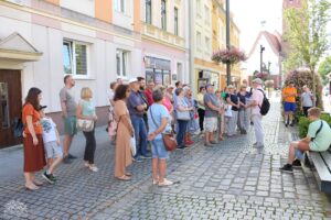 Grupa turystów z uwagą słucha swojego przewodnika na malowniczej ulicy w Polsce. Przewodnik, gestykulując, wskazuje na ciekawe detale architektoniczne, podczas gdy zaciekawieni słuchacze chłoną przekazywane informacje. Scena uchwycona w ciepłym, naturalnym świetle, podkreślająca atmosferę polskiego miasta i zaangażowanie grupy w poznawanie jego historii i kultury.