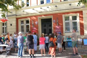 Grupa ludzi, wśród nich osoby z polską flagą, zgromadzona przed budynkiem. Na fasadzie budynku widnieje wyraźny napis "Rada Miejska w Oławie". Wszyscy słuchają z uwagą przewodnika, co sugeruje zorganizowane zwiedzanie lub wydarzenie.