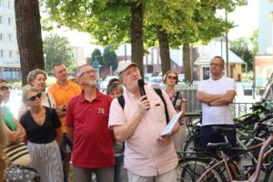 Mężczyzna z mikrofonem stoi przed grupą osób, przemawiając. W tle widoczne są drzewa i stojące rowery, co może wskazywać na plenerowe wydarzenie lub miejsce związane z aktywnością na świeżym powietrzu. Skupienie grupy na mówcy podkreśla charakter informacyjny lub prezentacyjny sceny.